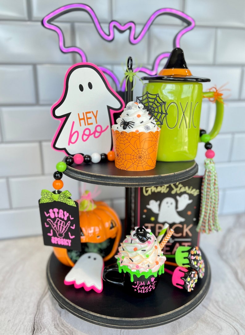 May include: A two-tiered black tiered tray decorated for Halloween. The top tier features a green mug with a witch's hat, a cupcake with a spider web and a spider, a ghost cutout that says "Hey Boo", and a beaded garland. The bottom tier features a black mug with green frosting and a ghost, a pumpkin, a sign that says "Stay Spooky", and a book that says "Ghost Stories Trick or Treat".