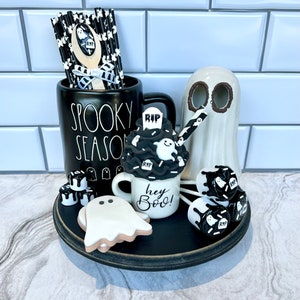 May include: A black and white Halloween themed tiered tray with a ghost mug that says "Hey Boo!" and a black mug that says "Spooky Season". The tray also has a ghost cookie, ghost-shaped marshmallows, and black and white striped straws.