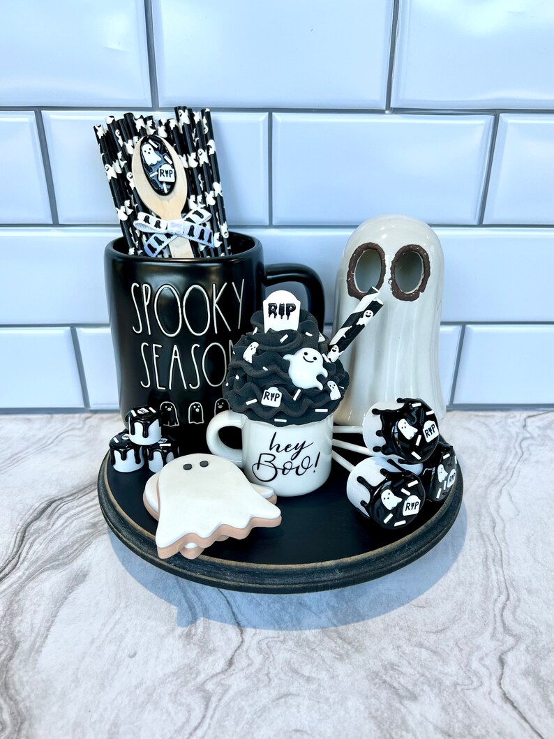 May include: A black and white Halloween themed tiered tray with a ghost mug that says "Hey Boo!" and a black mug that says "Spooky Season". The tray also has a ghost cookie, ghost-shaped marshmallows, and black and white striped straws.