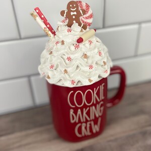 May include: Red ceramic mug with white lettering that reads "Cookie Baking Crew". The mug has a white whipped cream topping with a gingerbread man, candy cane, and a rolling pin on top.