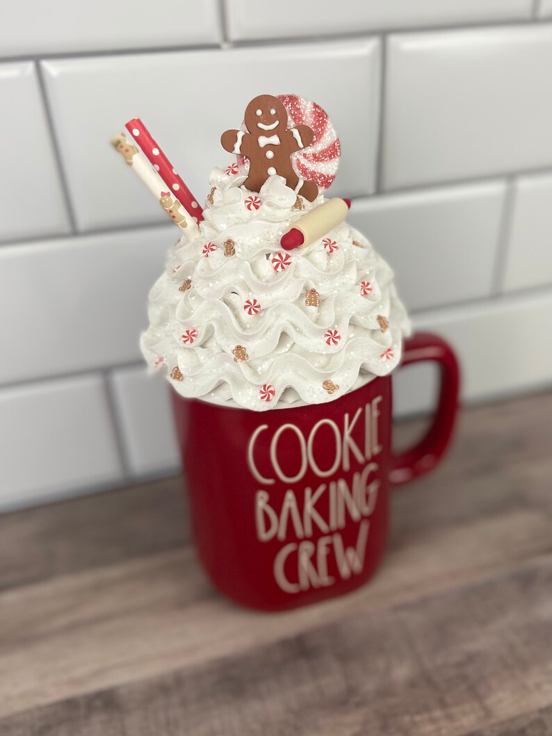 May include: Red ceramic mug with white lettering that reads "Cookie Baking Crew". The mug has a white whipped cream topping with a gingerbread man, candy cane, and a rolling pin on top.