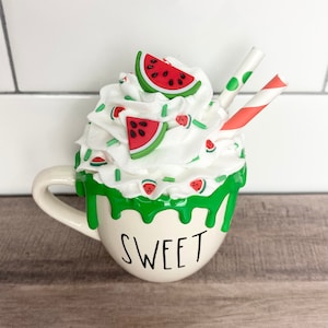 May include: White ceramic mug with the word "SWEET" on the front. The mug is filled with whipped cream, green sprinkles, and watermelon-shaped candies. Two red and white striped straws are sticking out of the whipped cream.