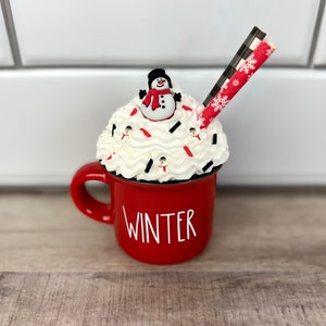 Op de afbeelding: Rode keramische mok met het woord "WINTER" aan de voorkant. De mok is gevuld met witte slagroom, bedekt met een sneeuwpopfiguur en rode en witte sprinkels. Een rood-zwarte gestreepte rietje steekt uit de slagroom.