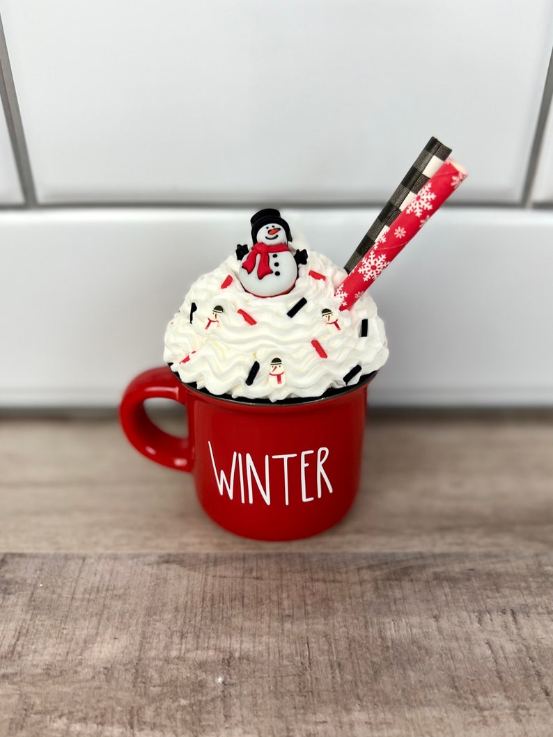 Op de afbeelding: Rode keramische mok met het woord "WINTER" aan de voorkant. De mok is gevuld met witte slagroom, bedekt met een sneeuwpopfiguur en rode en witte sprinkels. Een rood-zwarte gestreepte rietje steekt uit de slagroom.