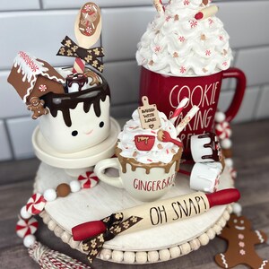 May include: A tiered tray decorated with a gingerbread theme. The tray features a white mug with a gingerbread man and the words "Baked with Love", a white mug with a gingerbread man and the words "Gingerbread", a red mug with the words "Cookie Baking", a rolling pin with the words "Oh Snap!" and a gingerbread man cookie. The tray is decorated with red and white candy canes and a white and red tassel.