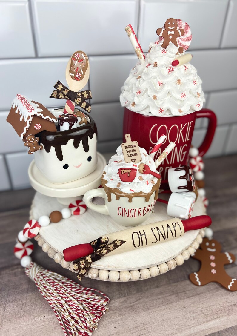 May include: A tiered tray decorated with a gingerbread theme. The tray features a white mug with a gingerbread man and the words "Baked with Love", a white mug with a gingerbread man and the words "Gingerbread", a red mug with the words "Cookie Baking", a rolling pin with the words "Oh Snap!" and a gingerbread man cookie. The tray is decorated with red and white candy canes and a white and red tassel.