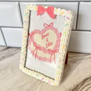 May include: White picture frame with a pink bow and sprinkles around the edge. The frame has a pink heart with the text "Bows & Silos" inside.