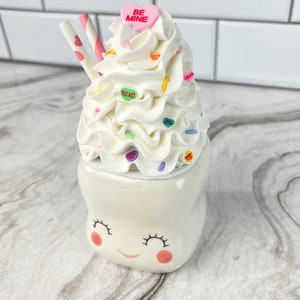 May include: White ceramic mug shaped like a smiling tooth with pink cheeks. The mug is topped with whipped cream, candy hearts, and two pink and white striped straws. The candy hearts have messages like "Be Mine", "Love You", and "XOXO".