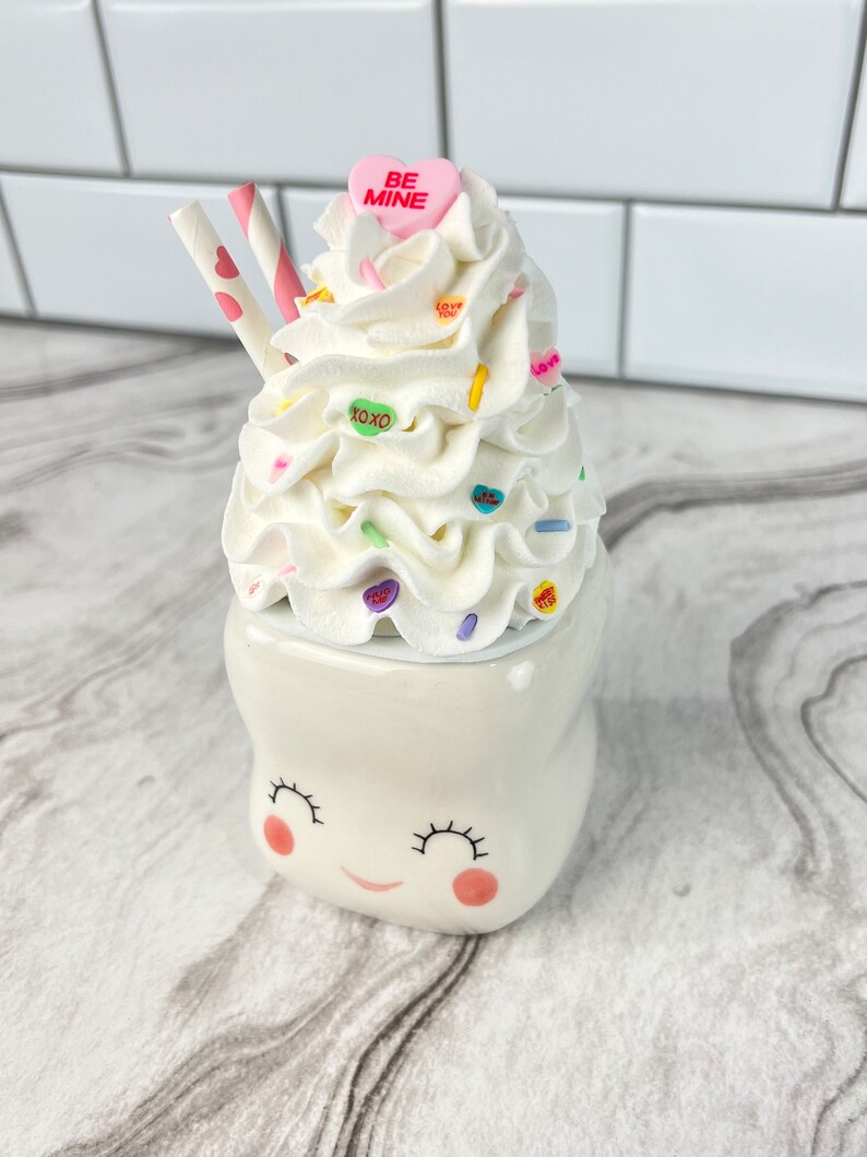 May include: White ceramic mug shaped like a smiling tooth with pink cheeks. The mug is topped with whipped cream, candy hearts, and two pink and white striped straws. The candy hearts have messages like "Be Mine", "Love You", and "XOXO".