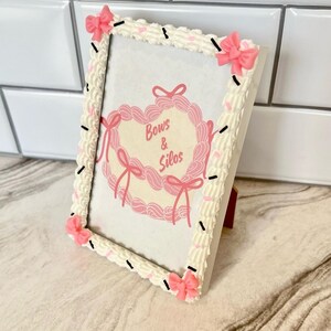 May include: White picture frame with a pink and white frosting design. The frame has pink bows on each corner. The frame has a picture inside with the text "Bows & Silos".