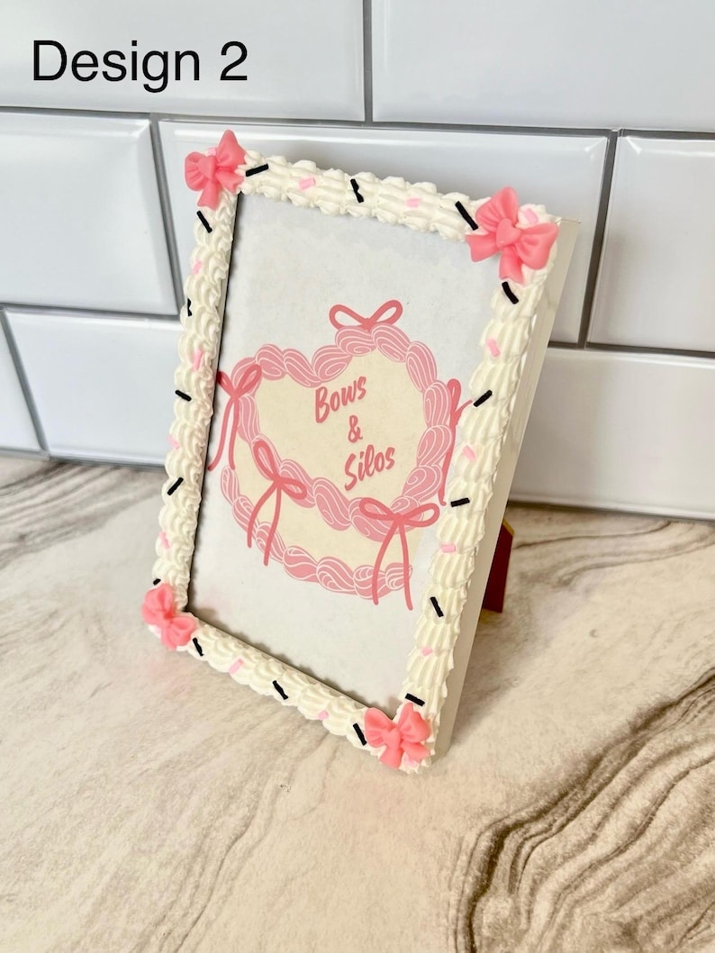May include: White picture frame with a pink and white frosting design. The frame has pink bows on each corner. The frame has a picture inside with the text "Bows & Silos".