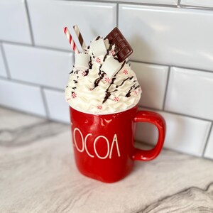 May include: Red ceramic mug with the word "COCOA" on the front. The mug is filled with whipped cream, chocolate sauce, marshmallows, and a chocolate bar. Two red and white striped straws are sticking out of the whipped cream.