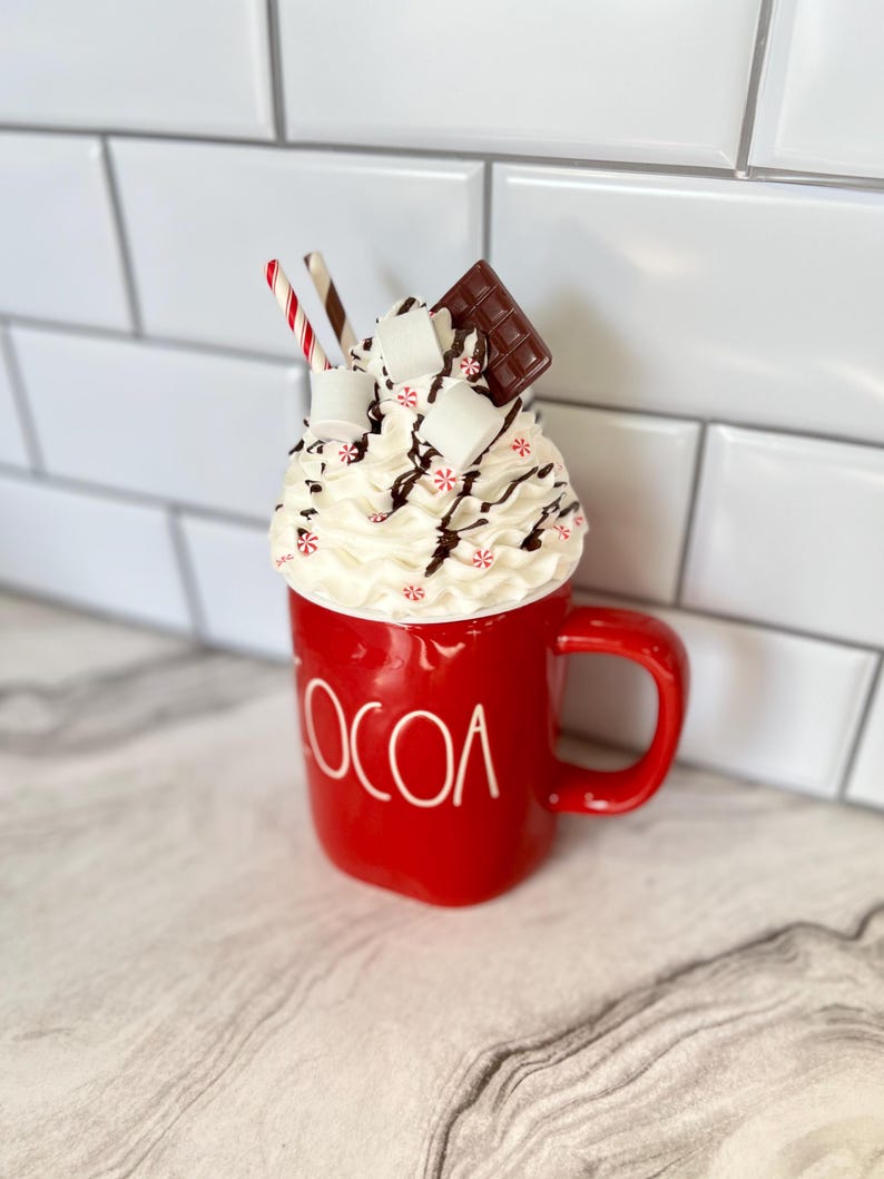 May include: Red ceramic mug with the word "COCOA" on the front. The mug is filled with whipped cream, chocolate sauce, marshmallows, and a chocolate bar. Two red and white striped straws are sticking out of the whipped cream.