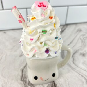 May include: A white ceramic mug with a smiling face, filled with white whipped cream and topped with colorful candy hearts. The mug has a straw and a pink candy heart with the text "BE MINE" on top.