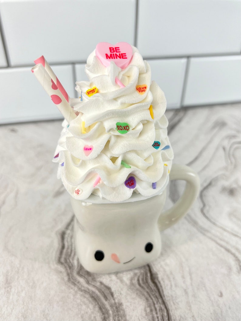 May include: A white ceramic mug with a smiling face, filled with white whipped cream and topped with colorful candy hearts. The mug has a straw and a pink candy heart with the text "BE MINE" on top.