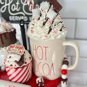 May include: A tiered serving tray with a red base and white trim. The top tier features a white mug with the words "HOT COCOA" in red lettering. The mug is filled with whipped cream, marshmallows, chocolate shavings, and a candy cane. The middle tier has a red and white striped cupcake with chocolate frosting, chocolate shavings, and a small sign that says "HOT COCOA". The bottom tier has a white rolling pin with the words "WARM UP HERE" in black lettering.