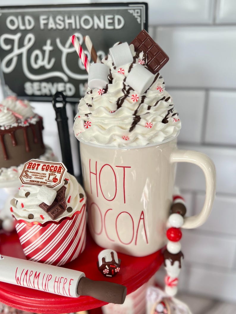 May include: A tiered serving tray with a red base and white trim. The top tier features a white mug with the words "HOT COCOA" in red lettering. The mug is filled with whipped cream, marshmallows, chocolate shavings, and a candy cane. The middle tier has a red and white striped cupcake with chocolate frosting, chocolate shavings, and a small sign that says "HOT COCOA". The bottom tier has a white rolling pin with the words "WARM UP HERE" in black lettering.