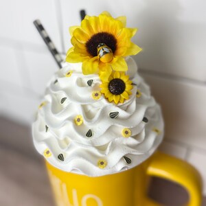 May include: Yellow ceramic mug with the text "Hello Sunshine" on the side. The mug is topped with white whipped cream and decorated with small yellow sunflowers and black sunflower seeds. A larger yellow sunflower with a small bee is on top of the whipped cream.