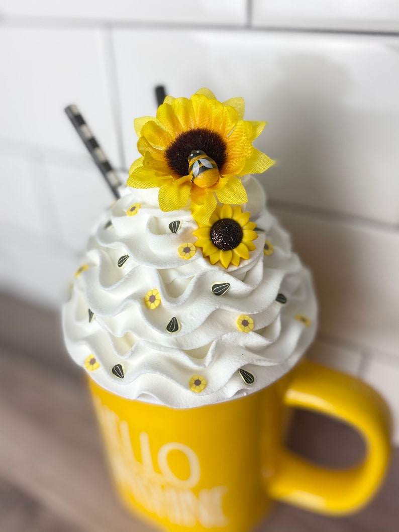 May include: Yellow ceramic mug with the text "Hello Sunshine" on the side. The mug is topped with white whipped cream and decorated with small yellow sunflowers and black sunflower seeds. A larger yellow sunflower with a small bee is on top of the whipped cream.