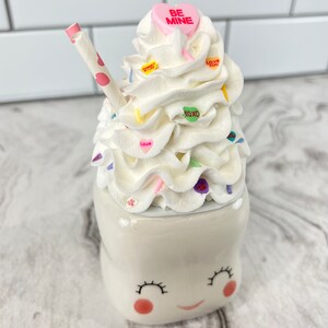 May include: A white ceramic mug shaped like a smiling face with pink cheeks and eyelashes. The mug is topped with whipped cream, colorful sprinkles, and candy hearts that say "Be Mine", "Love You", and "XOXO". A pink and white striped straw sticks out of the whipped cream.