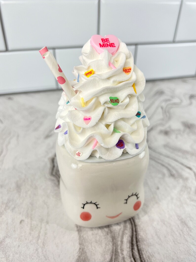 May include: A white ceramic mug shaped like a smiling face with pink cheeks and eyelashes. The mug is topped with whipped cream, colorful sprinkles, and candy hearts that say "Be Mine", "Love You", and "XOXO". A pink and white striped straw sticks out of the whipped cream.