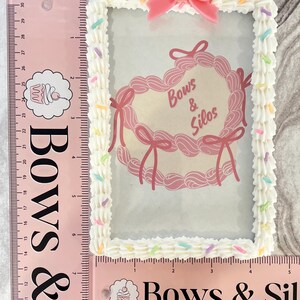 May include: A white picture frame with a pink heart-shaped design and the text "Bows & Silos". The frame is decorated with white frosting and colorful sprinkles.