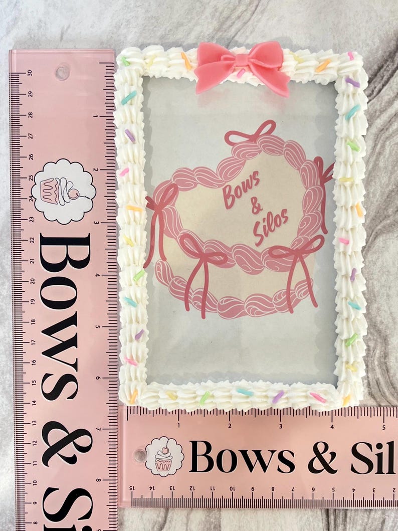 May include: A white picture frame with a pink heart-shaped design and the text "Bows & Silos". The frame is decorated with white frosting and colorful sprinkles.