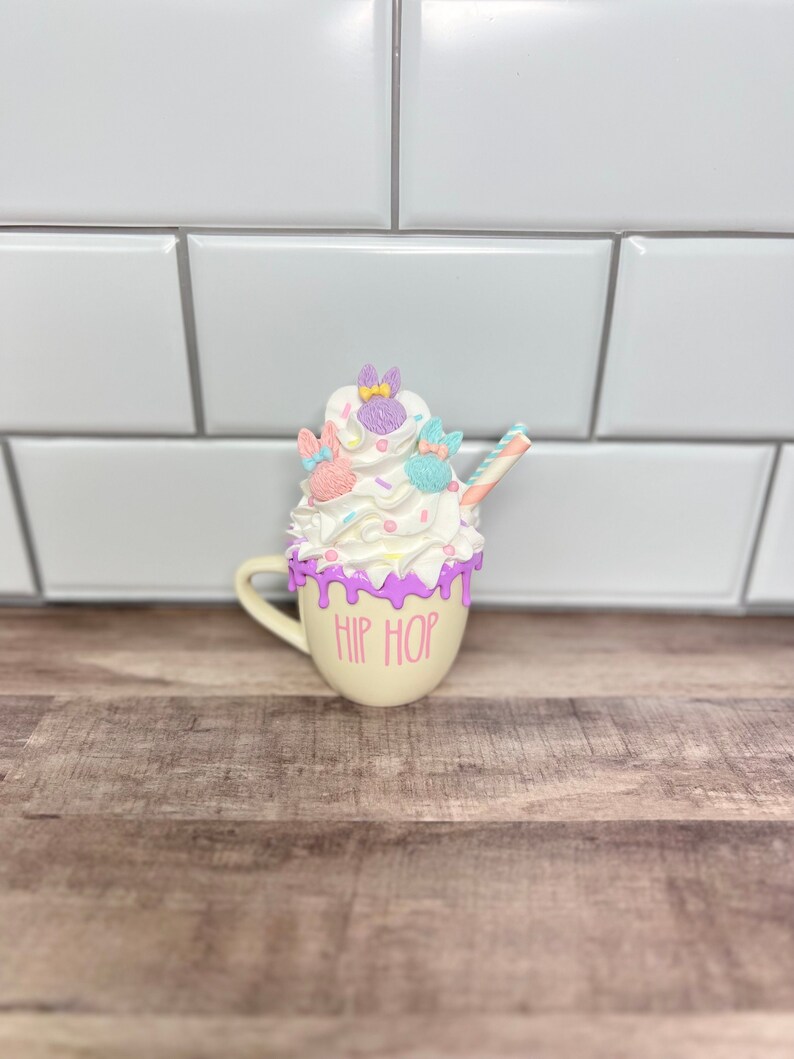 K&ouml;nnte beinhalten: Eine wei&szlig;e Keramiktasse mit rosa und lila Glasur und einer Sahnehaube mit pastellfarbenen Hasendekorationen. Die Tasse tr&auml;gt den Schriftzug "HIP HOP".
