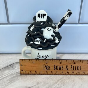 May include: A white ceramic mug with a black and white ghost and tombstone design. The mug has the text "hey boo" written on it. The mug is decorated with black and white frosting and a black and white striped straw.