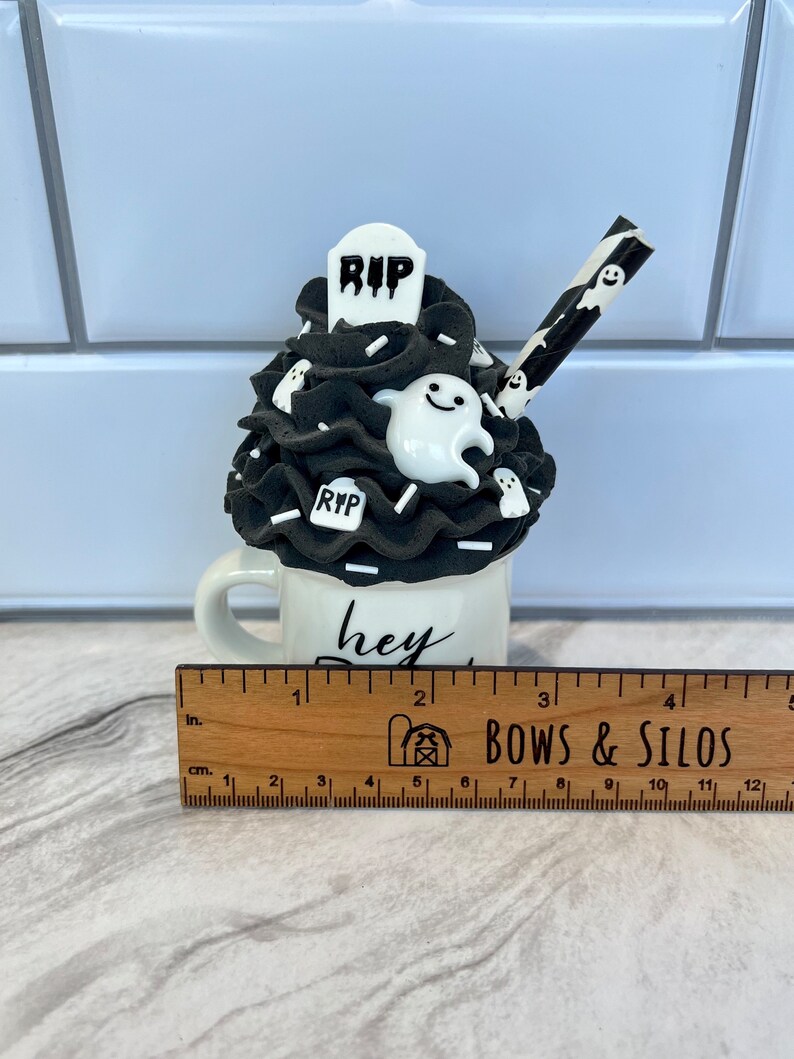 May include: A white ceramic mug with a black and white ghost and tombstone design. The mug has the text "hey boo" written on it. The mug is decorated with black and white frosting and a black and white striped straw.