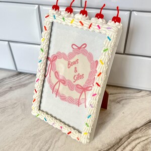 May include: White picture frame with a cake frosting design, featuring red cherries and colorful sprinkles. The frame has a pink heart-shaped image inside with the text "Bows & Silos".