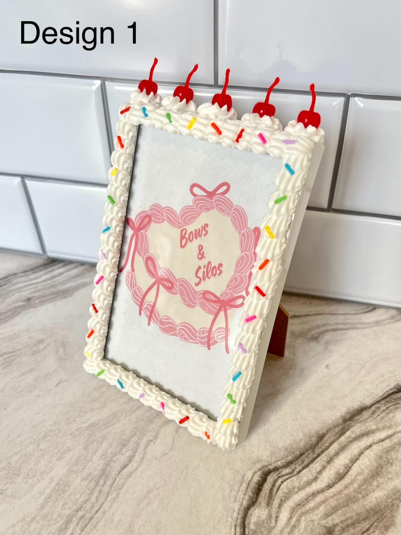 May include: White picture frame with a cake frosting design, featuring red cherries and colorful sprinkles. The frame has a pink heart-shaped image inside with the text "Bows & Silos".