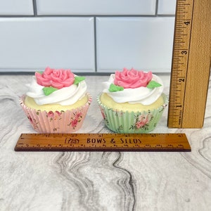 May include: Two cupcakes decorated with pink roses and green leaves. The cupcakes are in pink and green floral cupcake liners. A ruler is in the image for size reference.