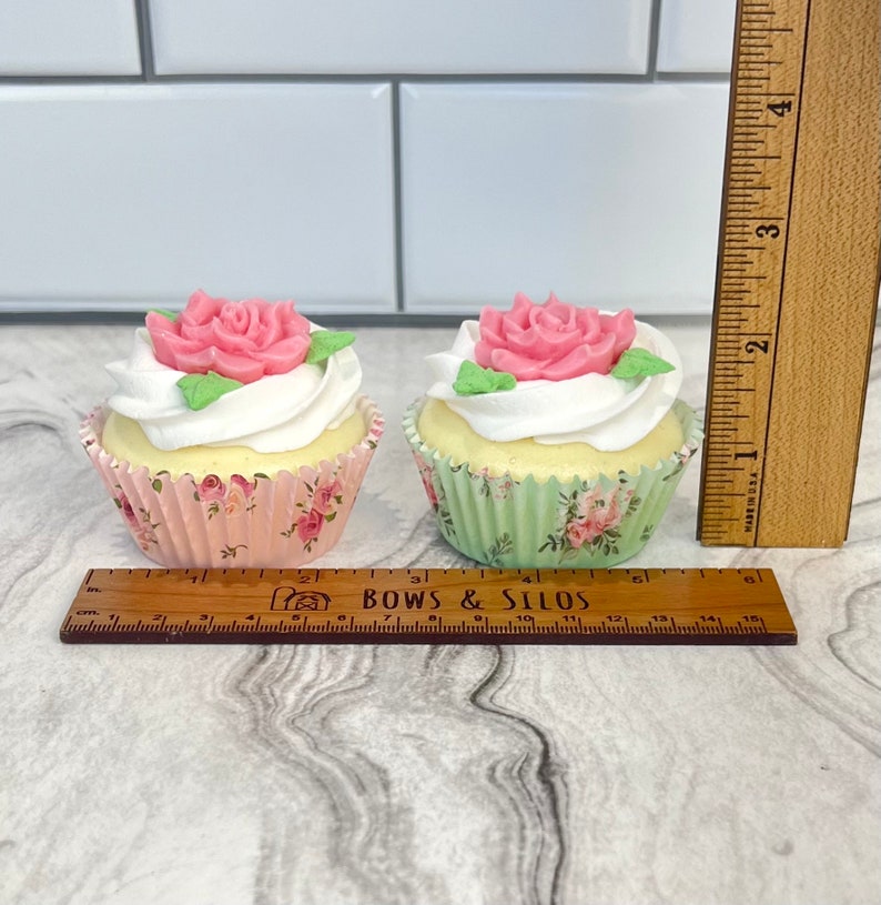 May include: Two cupcakes decorated with pink roses and green leaves. The cupcakes are in pink and green floral cupcake liners. A ruler is in the image for size reference.