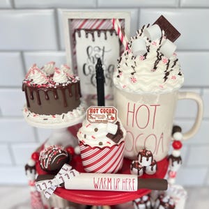 May include: A three-tiered serving tray with a red base and white trim. The top tier features a white mug with red lettering that says "HOT COCOA". The mug is filled with white whipped cream, chocolate shavings, and red and white candy canes. The middle tier features a chocolate cake with chocolate frosting and red and white candy cane sprinkles. The bottom tier features a cupcake with red and white striped paper and a chocolate cupcake with white frosting and chocolate shavings. The tray also includes a rolling pin with the words "WARM UP HERE" written on it.