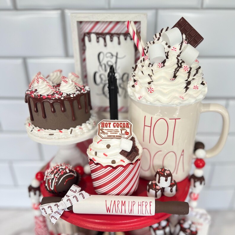 May include: A three-tiered serving tray with a red base and white trim. The top tier features a white mug with red lettering that says "HOT COCOA". The mug is filled with white whipped cream, chocolate shavings, and red and white candy canes. The middle tier features a chocolate cake with chocolate frosting and red and white candy cane sprinkles. The bottom tier features a cupcake with red and white striped paper and a chocolate cupcake with white frosting and chocolate shavings. The tray also includes a rolling pin with the words "WARM UP HERE" written on it.