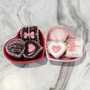 May include: Two heart-shaped bowls filled with assorted chocolates. The red bowl contains three chocolates: a dark chocolate with white stripes, a dark chocolate with a pink heart, and a dark chocolate with a pink bow. The pink bowl contains three chocolates: a white chocolate with pink stripes, a white chocolate with a pink heart, and a white chocolate with a pink bow.