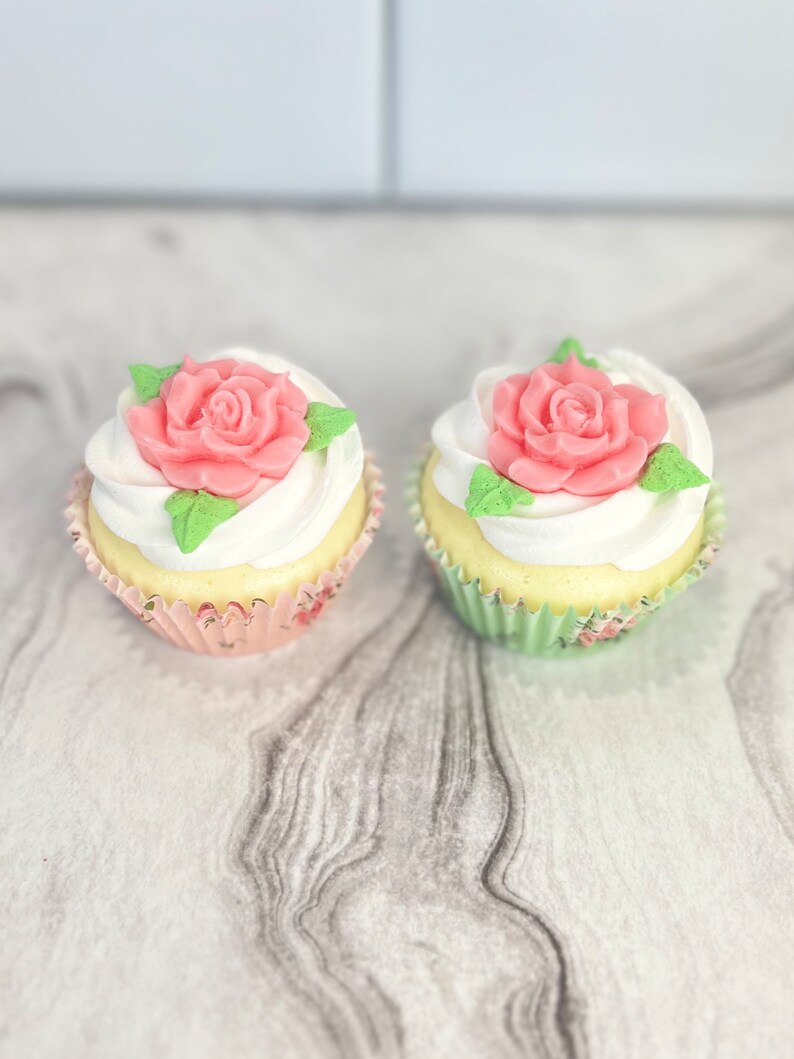 May include: Two cupcakes with white frosting and pink rose decorations. The cupcakes are in paper liners with floral patterns.