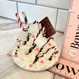 May include: A white chocolate-covered marshmallow treat with chocolate sauce, candy canes, and a chocolate bar on top. The treat is on a white plate and is about 10 cm tall.