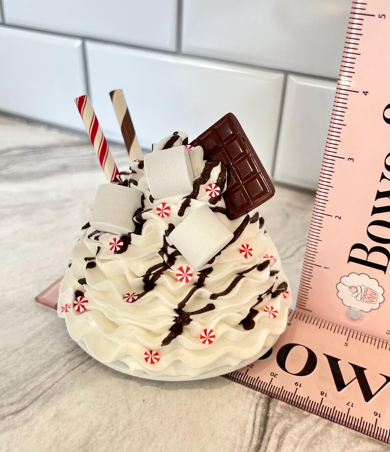 May include: A white chocolate-covered marshmallow treat with chocolate sauce, candy canes, and a chocolate bar on top. The treat is on a white plate and is about 10 cm tall.