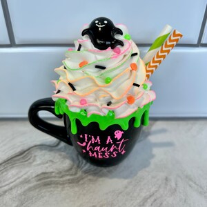 May include: Black coffee mug with green slime dripping down the side. The mug has a white whipped cream topping with sprinkles, a ghost figurine, and a striped straw. The mug reads "I'm a haunt mess".