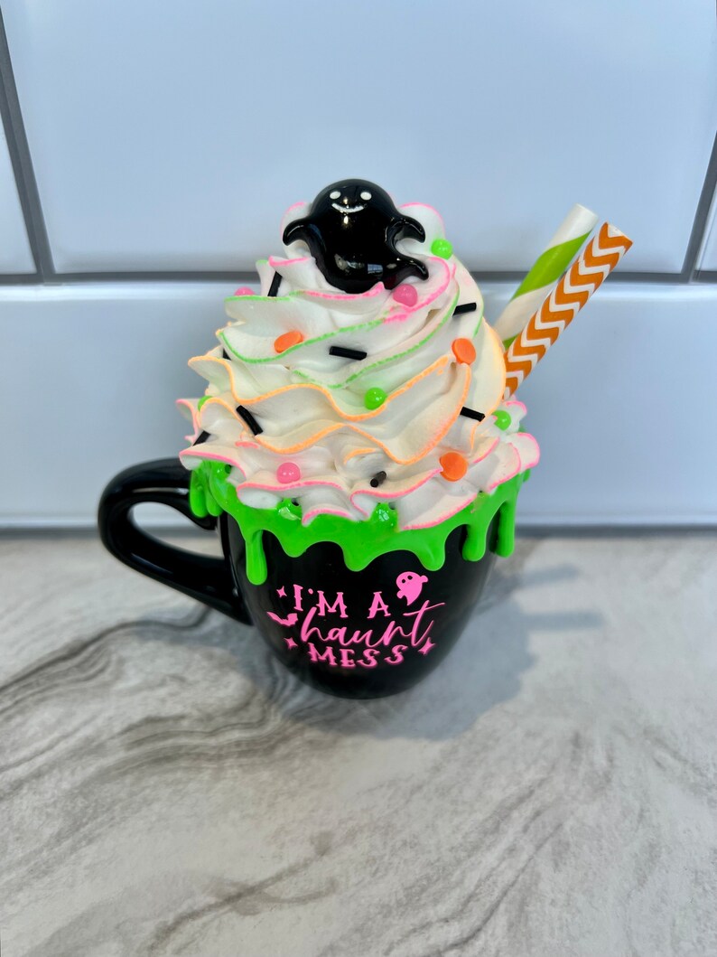 May include: Black coffee mug with green slime dripping down the side. The mug has a white whipped cream topping with sprinkles, a ghost figurine, and a striped straw. The mug reads "I'm a haunt mess".