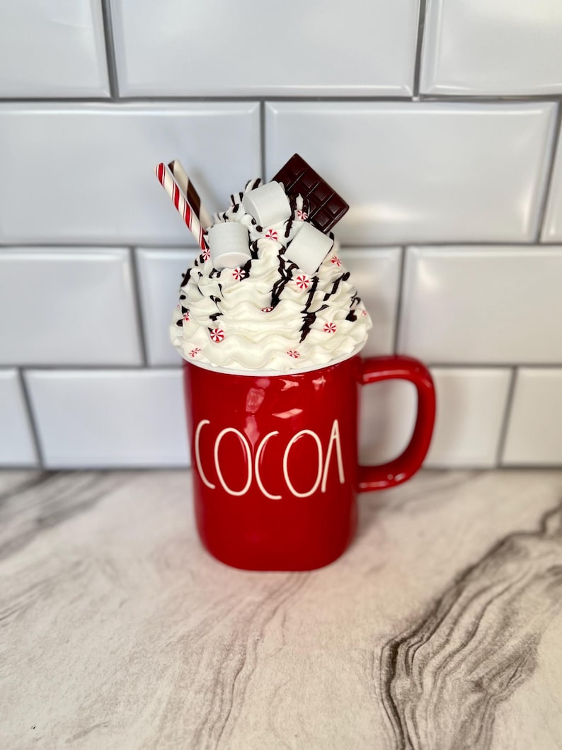 May include: Red ceramic mug with a white "COCOA" inscription. The mug is filled with whipped cream, marshmallows, chocolate pieces, and a red and white candy cane.
