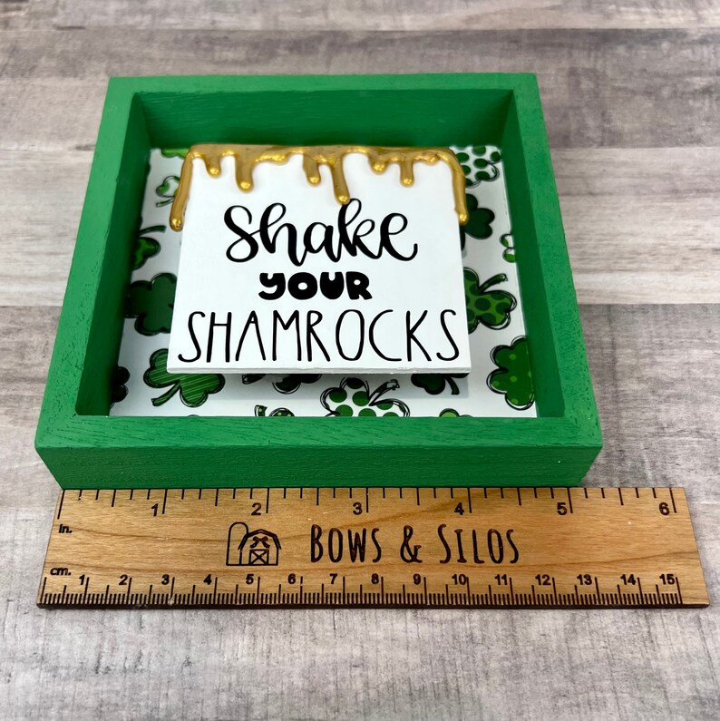 以下が含まれることがあります： 緑色の木製のフレームの中に、「Shake Your Shamrocks」と書かれた白い看板が入っています。看板には金色の塗料が前面に垂れており、フレームの背景は緑色のクローバー柄です。