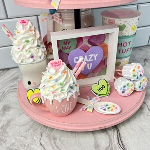 May include: A pink tiered tray decorated for Valentine's Day with a hot chocolate mug, a milkshake, and a framed picture with the words "Crazy U". The tray is decorated with candy hearts, sprinkles, and other Valentine's Day themed decorations.