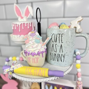 K&ouml;nnte beinhalten: Ein dreistufiges Tablett, das f&uuml;r Ostern mit einem rosa Hasenkuchen, einer wei&szlig;en Tasse mit dem Text "Cute as a Bunny", einer wei&szlig;en Tasse mit dem Text "Hip Hop", einem gelben Nudelholz mit dem Text "Egg Hunt" und einer Schnur pastellfarbener Perlen dekoriert ist.