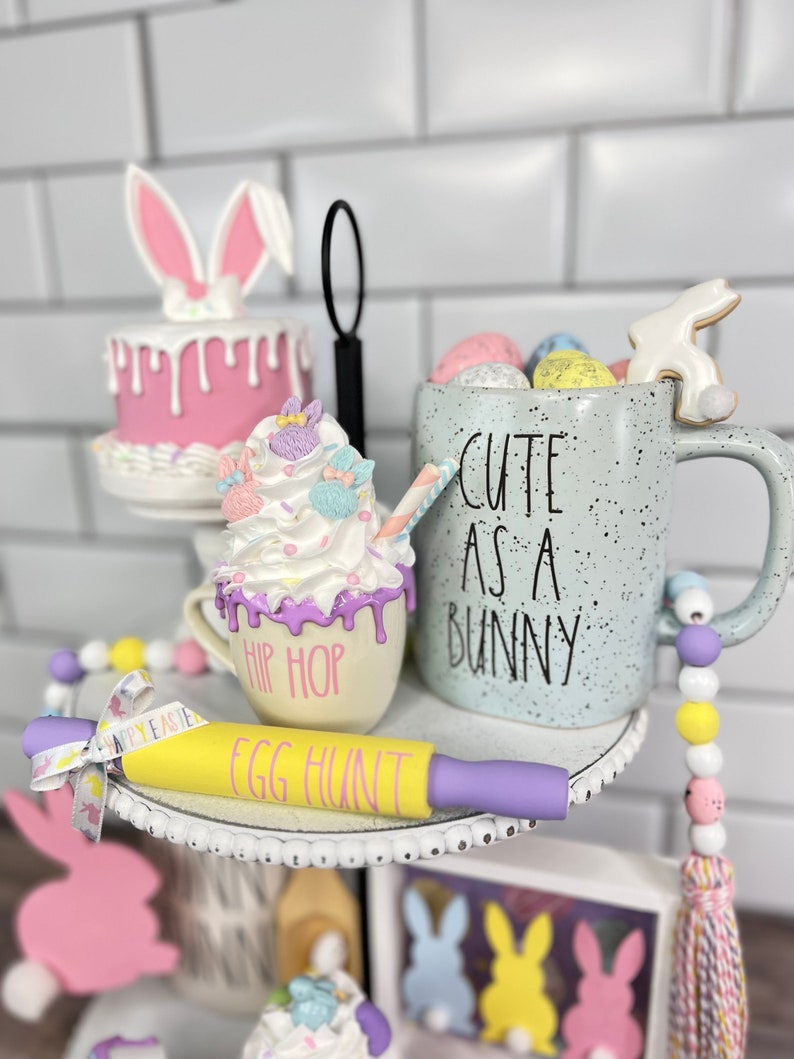K&ouml;nnte beinhalten: Ein dreistufiges Tablett, das f&uuml;r Ostern mit einem rosa Hasenkuchen, einer wei&szlig;en Tasse mit dem Text "Cute as a Bunny", einer wei&szlig;en Tasse mit dem Text "Hip Hop", einem gelben Nudelholz mit dem Text "Egg Hunt" und einer Schnur pastellfarbener Perlen dekoriert ist.