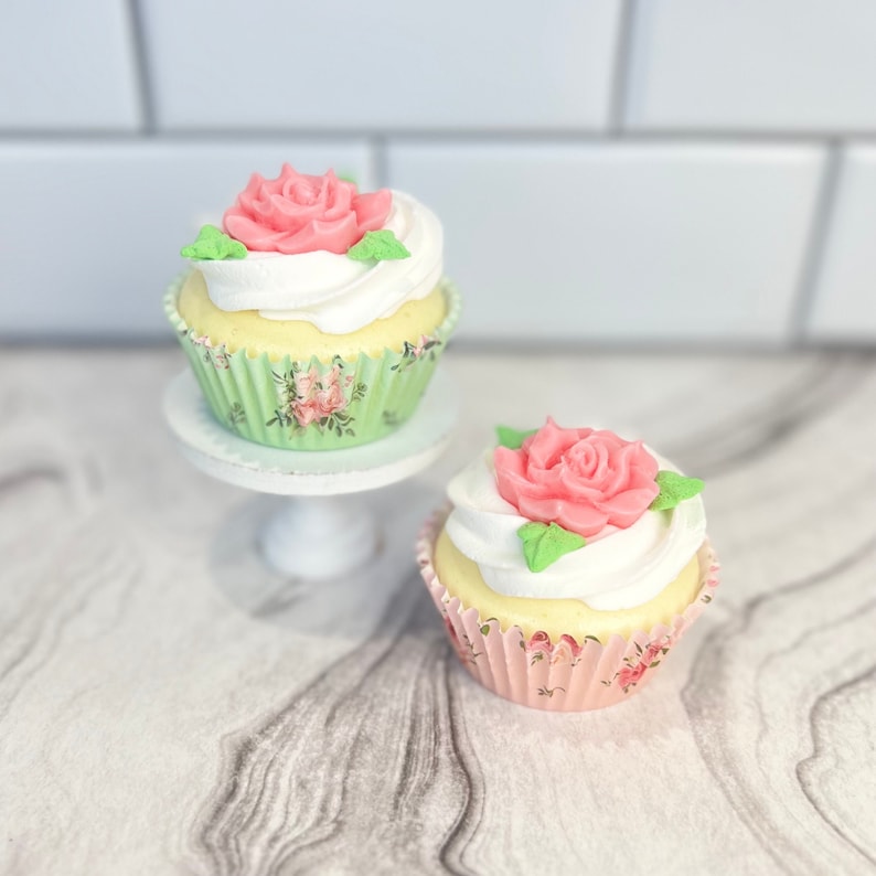 May include: Two cupcakes with white frosting and pink rose decorations. The cupcakes are in floral patterned paper liners. One cupcake is on a white cake stand.