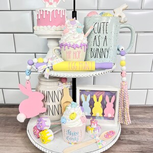 K&ouml;nnte beinhalten: Ein zweistufiges wei&szlig;es Tablett mit Etageren, das mit Ostern-Motiven dekoriert ist. Die obere Etage zeigt einen rosa Kuchen mit Hasenohren, eine pastellfarbene Tasse mit dem Text "Cute as a Bunny" und einen gelben Nudelholz mit dem Text "Hip Hop Egg Hunt". Die untere Etage zeigt eine rosa Hasenausschneidung, einen Cupcake mit pastellfarbenen Streuseln und einen Holzl&ouml;ffel. Das Tablett ist mit wei&szlig;en Perlen und einem wei&szlig;en Band verziert.
