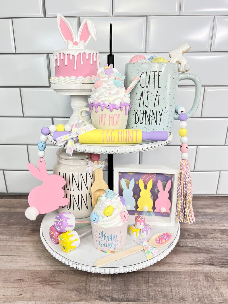 K&ouml;nnte beinhalten: Ein zweistufiges wei&szlig;es Tablett mit Etageren, das mit Ostern-Motiven dekoriert ist. Die obere Etage zeigt einen rosa Kuchen mit Hasenohren, eine pastellfarbene Tasse mit dem Text "Cute as a Bunny" und einen gelben Nudelholz mit dem Text "Hip Hop Egg Hunt". Die untere Etage zeigt eine rosa Hasenausschneidung, einen Cupcake mit pastellfarbenen Streuseln und einen Holzl&ouml;ffel. Das Tablett ist mit wei&szlig;en Perlen und einem wei&szlig;en Band verziert.